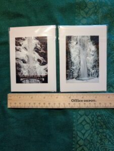 Aerial photo looking down at the front side of two small photo notecards featuring upper Multnomah Falls frozen (including the Benson bridge) on the left, and the lower falls frozen on the right. A wooden ruler shows beneath for scale.