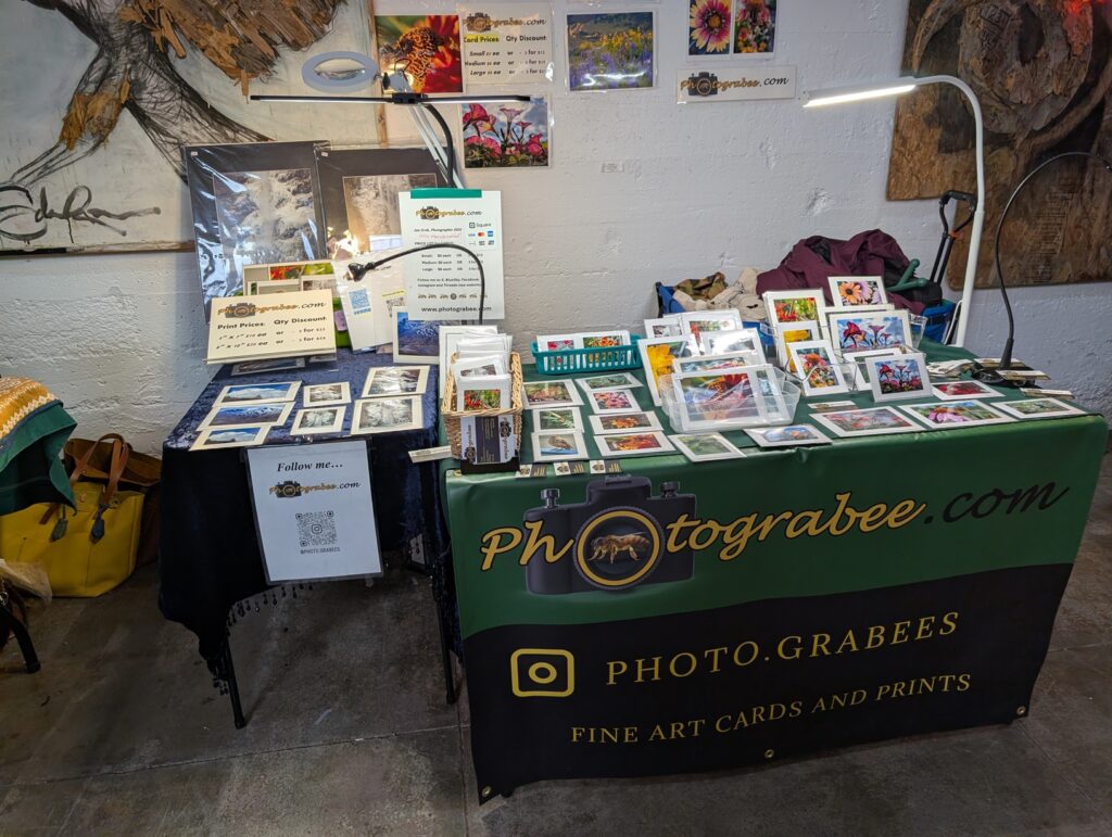 Photo showing two small tables set up at 90 degree angle to each other against a white wall with art hung on it. Banner on front reads Photograbee.com and has an insta address. Many colorful photo notecards of bees and flowers and mountains adorn the top of the tables.