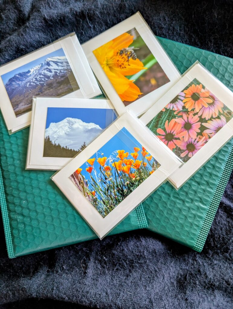 Aerial shot looking down on an array of five of Jan's small colorful photo notecards featuring clockwise from top left: Mt St Helens up close, a honeybee in a golden choriopsis blossom in the sun, a colorful spray of pink and salmon cone flowers, a joyful shot from a little beneath a patch of bright orange California poppies reaching for the blue sky, and a bright white shot of a very snow-covered Mt Hood with a dark green tree line coming in from the left at the bottom against a blue sky. These are set on a couple of emerald green plastic mailers on top of a dark blue velvet blanket for contrast.