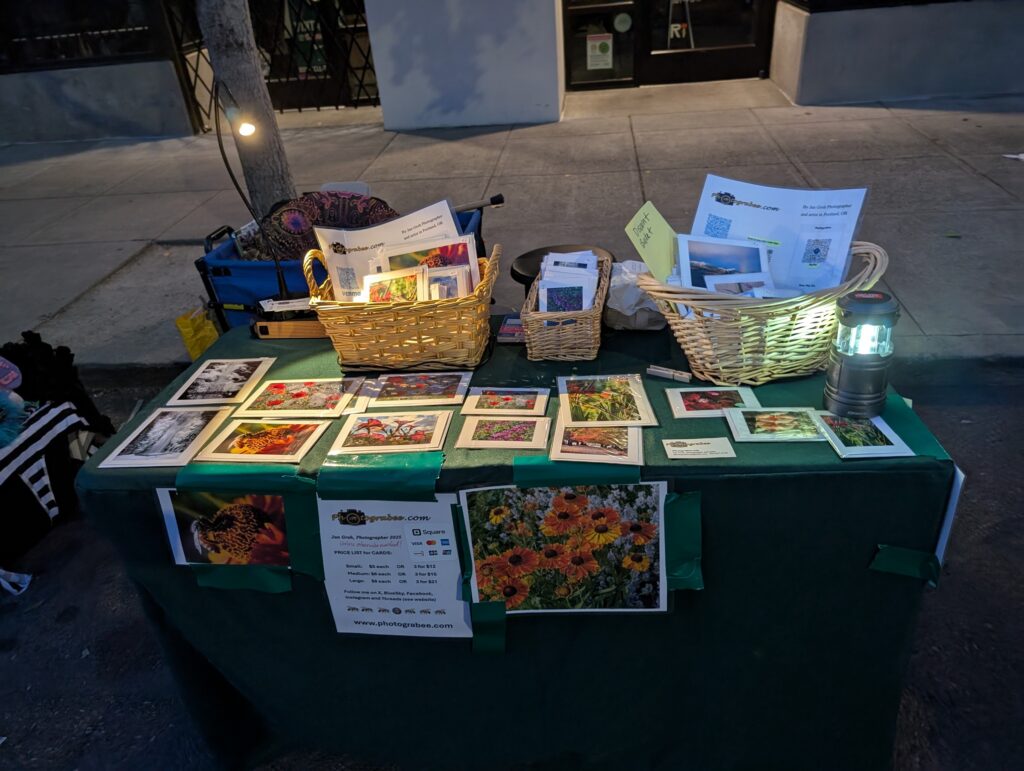 A humble 2x4' table covered in emerald green cloth with Jan's colorful photo notecards displayed flat and in baskets. Some battery powered lighting helps to see them as dusk sets in. North side of NE Alberta Street August 2025.
